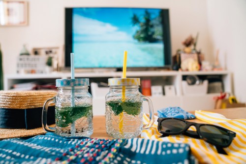 Two mason jars with mint drinks and colorful straws sit on a table, with a beach scene on the TV and summer accessories nearby.