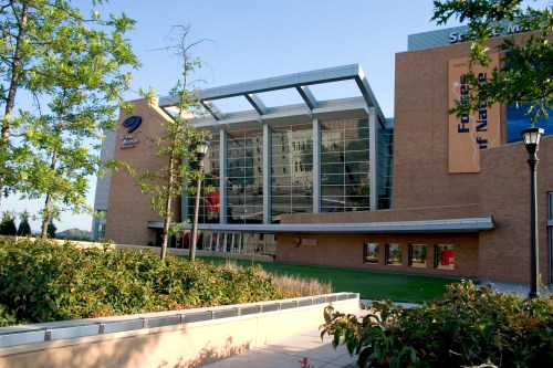 A modern building with large glass windows, surrounded by greenery and trees, showcasing a bright blue sky.