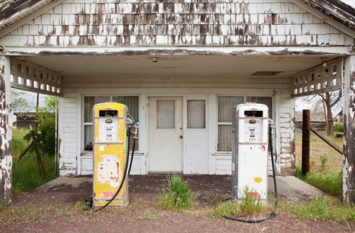 Abandoned gas station with two vintage pumps in front of a weathered white building. Overgrown grass surrounds the area.