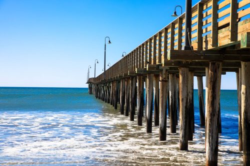 A wooden pier extends over calm blue waters, with waves gently lapping at its supports under a clear sky.