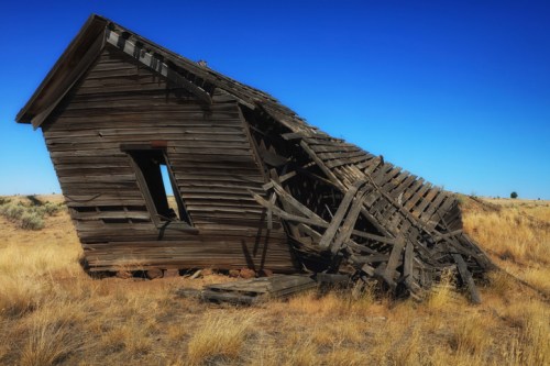 A dilapidated wooden house leaning to one side, surrounded by dry grass under a clear blue sky.