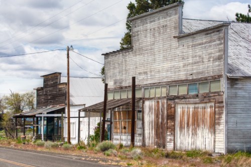 Abandoned wooden storefronts line a quiet road, with weathered facades and overgrown vegetation under a cloudy sky.