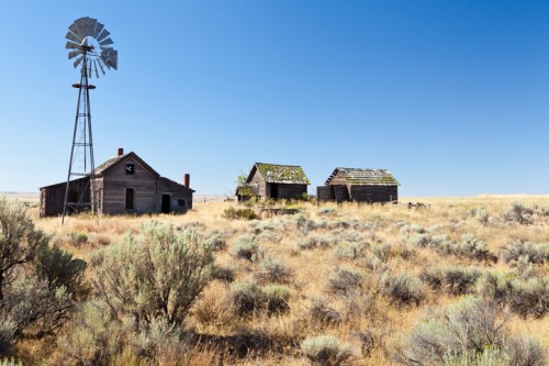 Abandoned buildings and a windmill in a dry, grassy landscape under a clear blue sky.