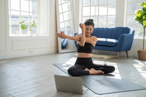 A woman in activewear stretches while sitting on a yoga mat in a bright, modern room with a laptop in front of her.