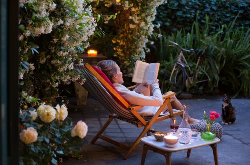 A woman relaxes in a chair, reading a book in a garden surrounded by flowers, candles, and a cat nearby.