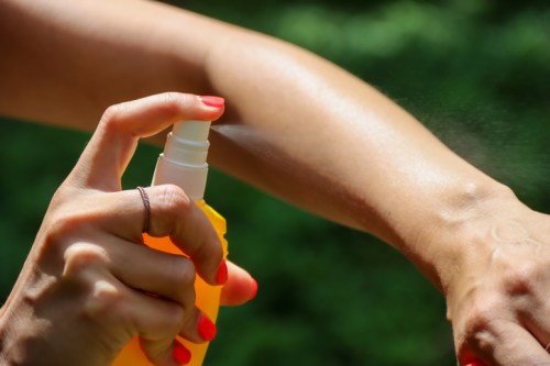A hand sprays sunscreen onto an arm, with a blurred green background.