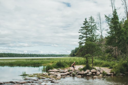 A person walks along a rocky shore by a calm lake, surrounded by lush greenery and cloudy skies.
