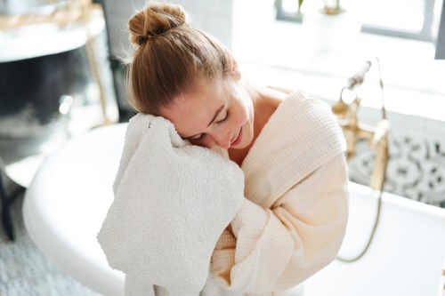 A woman in a bathrobe gently hugs a towel, smiling softly in a bright bathroom with a bathtub in the background.
