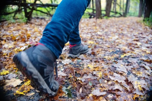 A close-up of a person's hiking boots walking on a leaf-covered forest trail in autumn.