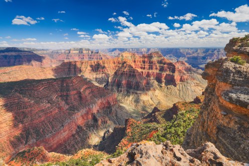 A panoramic view of the Grand Canyon, showcasing colorful rock layers and a clear blue sky with scattered clouds.
