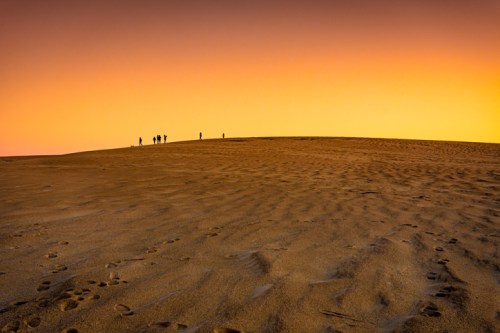 Silhouetted figures walking on a sandy dune at sunset, with a gradient sky transitioning from orange to purple.