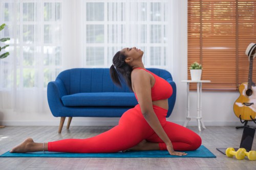 A woman in a red workout outfit performs a yoga pose on a mat in a bright, modern living room.