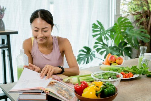 A woman in a tank top reads a cookbook at a table filled with fresh vegetables and fruits.