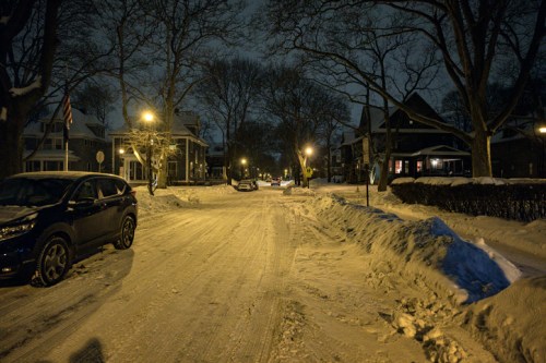 A snowy street at night, lined with houses and streetlights illuminating the scene.