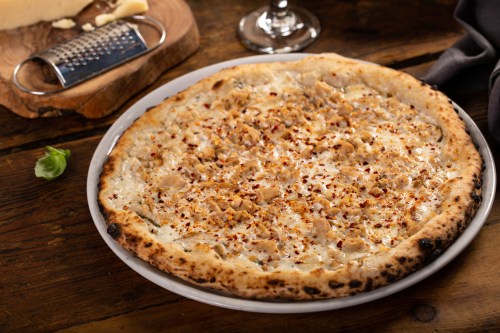 A freshly baked pizza topped with creamy sauce, chicken, and crushed red pepper flakes on a wooden table.