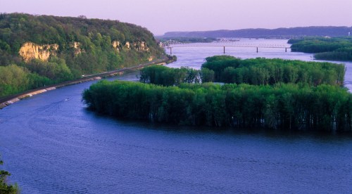 A serene river winding through lush greenery, with cliffs in the background and a bridge in the distance under a twilight sky.