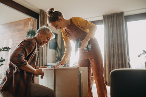A man and woman work together to assemble furniture, smiling and using a power drill in a bright, cozy room.