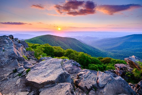 Sunset over rolling green mountains, with rocky foreground and colorful sky filled with clouds.