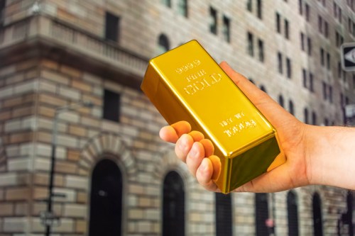 A hand holds a large gold bar in front of a historic building.