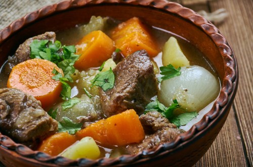 A bowl of hearty beef stew with carrots, potatoes, and herbs, served in a rustic clay dish.