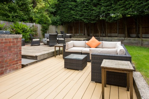 A cozy outdoor patio with a sectional sofa, orange cushion, glass table, and wooden deck surrounded by greenery.