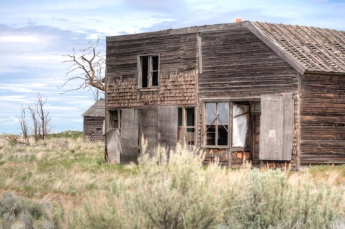 Abandoned wooden house with broken windows, surrounded by tall grass and a cloudy sky.
