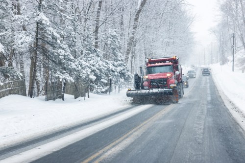 A snowplow clears a snowy road lined with trees, while cars navigate the winter weather.