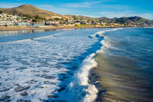 A scenic beach view with gentle waves lapping at the shore, framed by hills and coastal homes under a clear blue sky.