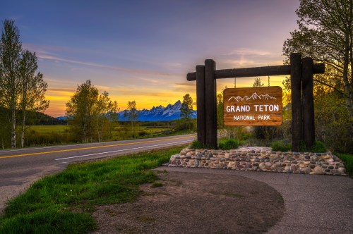 Sign for Grand Teton National Park at sunset, with mountains in the background and a scenic road in the foreground.