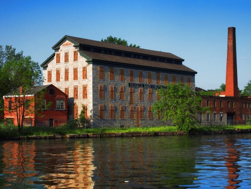 Historic industrial building by a river, featuring brick and stone architecture, with a tall chimney and lush greenery.