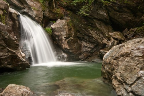 A serene waterfall cascades over rocks into a tranquil pool, surrounded by lush greenery and rugged stone formations.