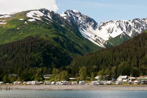 A scenic view of snow-capped mountains behind a line of RVs along a tranquil waterfront. Lush greenery surrounds the area.