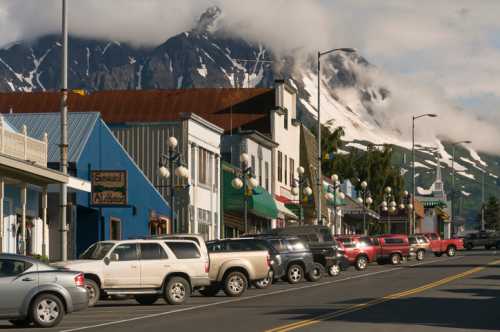 A scenic view of a small town street lined with parked cars, framed by mountains and cloudy skies in the background.