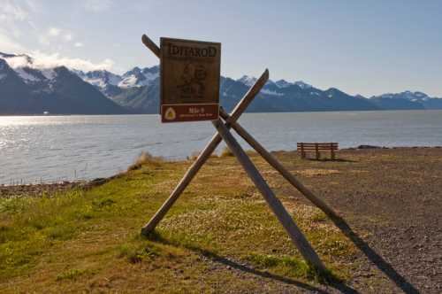 Sign marking the start of the Iditarod Trail, with mountains and water in the background. Bench nearby.