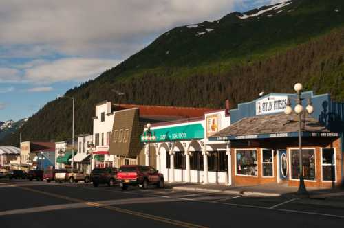 A scenic view of a small town street with colorful buildings, parked cars, and mountains in the background.