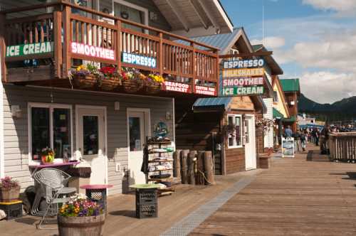 Colorful storefronts line a wooden boardwalk, featuring signs for ice cream, smoothies, and seafood. Bright flowers adorn the scene.