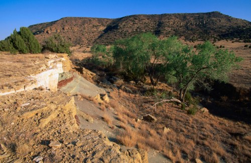 A dry landscape with rocky terrain, sparse vegetation, and a distant hill under a clear blue sky.