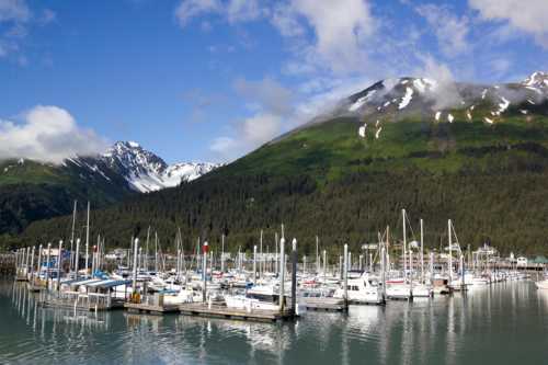 A scenic marina with boats docked, surrounded by lush green mountains and a partly cloudy blue sky.