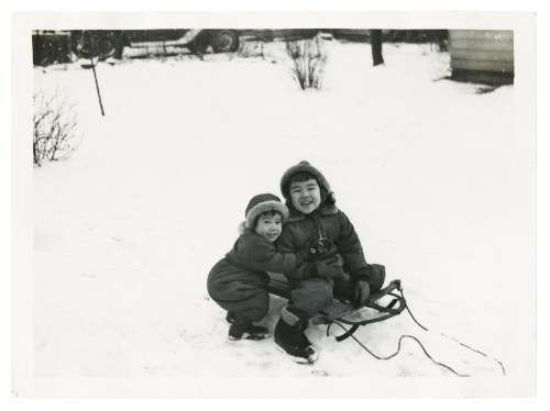 Two children in winter clothing smile while hugging a sled in a snowy outdoor setting.
