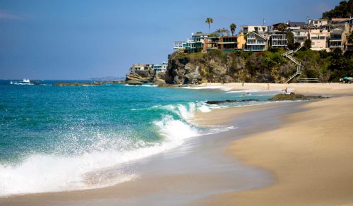 A serene beach scene with gentle waves, rocky cliffs, and coastal homes under a clear blue sky.