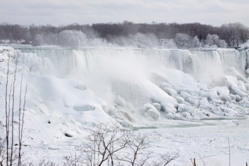A frozen waterfall with icy mist and snow-covered rocks, surrounded by bare trees and a cloudy sky.