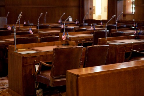 Empty legislative chamber with wooden desks, microphones, and small American flags on each desk.