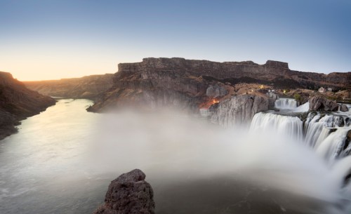 A scenic view of a waterfall cascading into a river, surrounded by rocky cliffs and a misty atmosphere at sunset.