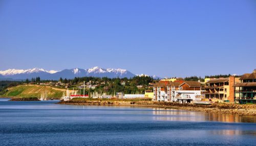 Scenic waterfront view with mountains in the background, featuring buildings along the shore and clear blue skies.