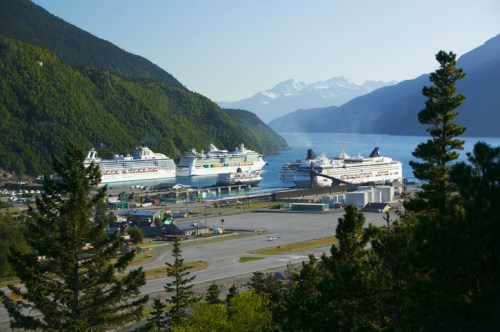 A scenic view of a harbor with multiple cruise ships docked, surrounded by mountains and lush greenery.