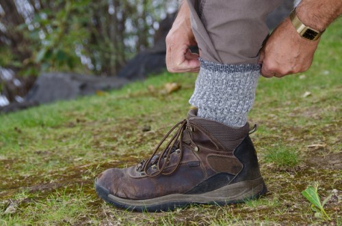 A person adjusts their gray sock above a brown hiking boot while standing on grassy ground.