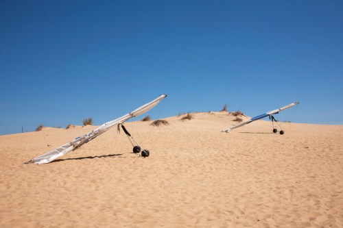 Two hang gliders resting on a sandy dune under a clear blue sky.