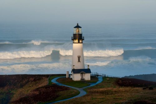 A lighthouse stands on a cliff, surrounded by crashing waves and a misty ocean backdrop.