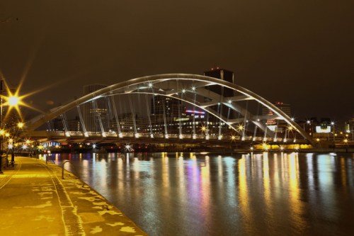 A illuminated bridge arches over a river at night, reflecting city lights and buildings in the water below.