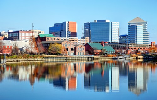 City skyline reflecting on a calm river, featuring modern buildings and colorful autumn foliage along the shore.